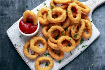 A pile of golden battered onion rings on a white wooden board, sprinkled with parsley, with a small bowl of ketchup