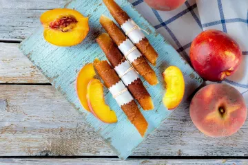 Rolled strips of peach fruit leather tied with paper and string on a pale-blue board, with fresh whole peaches, a halved peach and slices