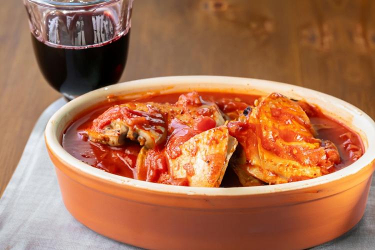 A small clay pot with chicken in tomato sauce and a glass of red wine.