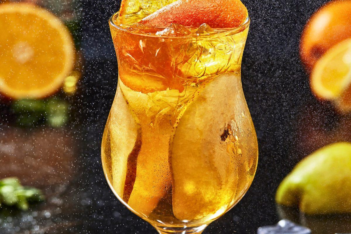 Cider spritz cocktail with ice and slices of orange in a curved glass against a misty, festive background.