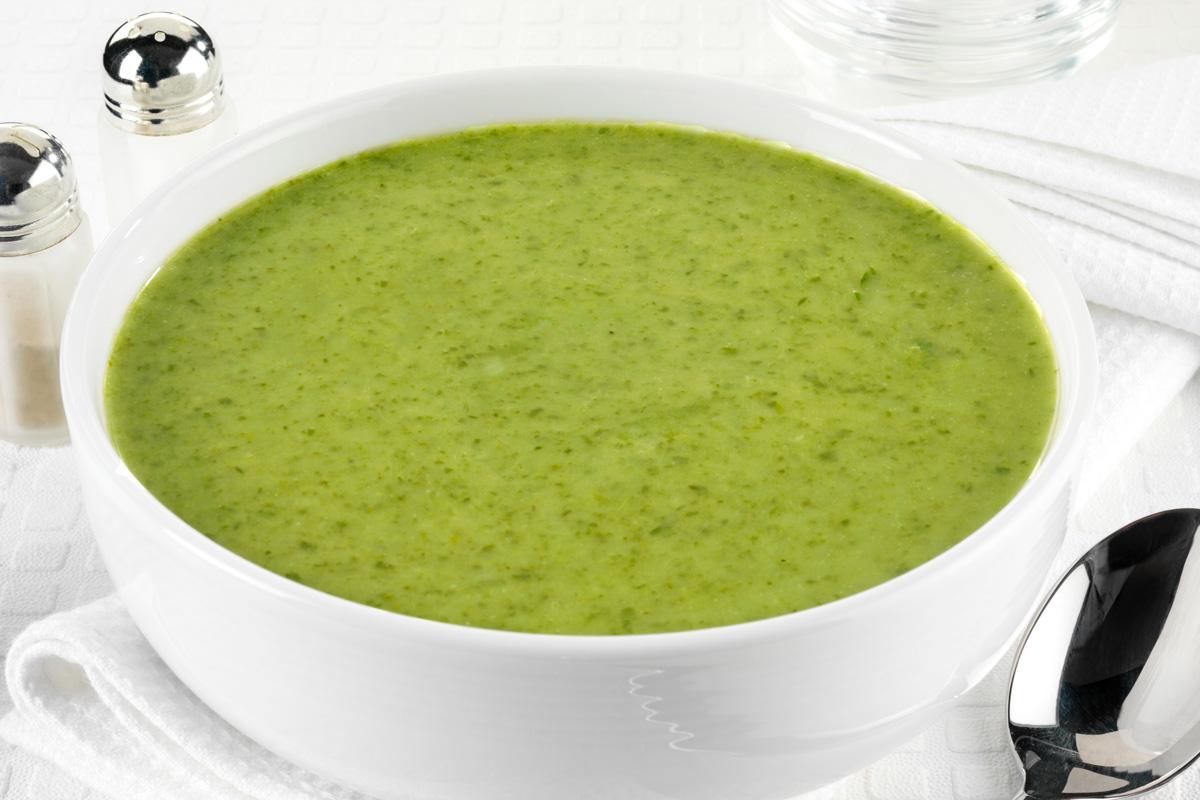A white bowl filled with chilled watercress soup on a white tablecloth, accompanied by a folded white napkin, a spoon, and salt and pepper shakers in the background.