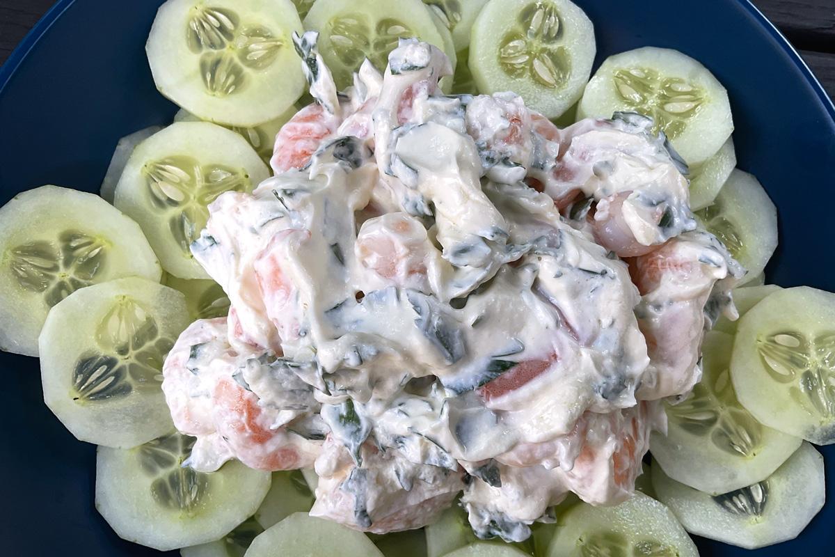 A prawn salad with creamy dressing and herbs, surrounded by neatly arranged cucumber slices on a dark blue plate.