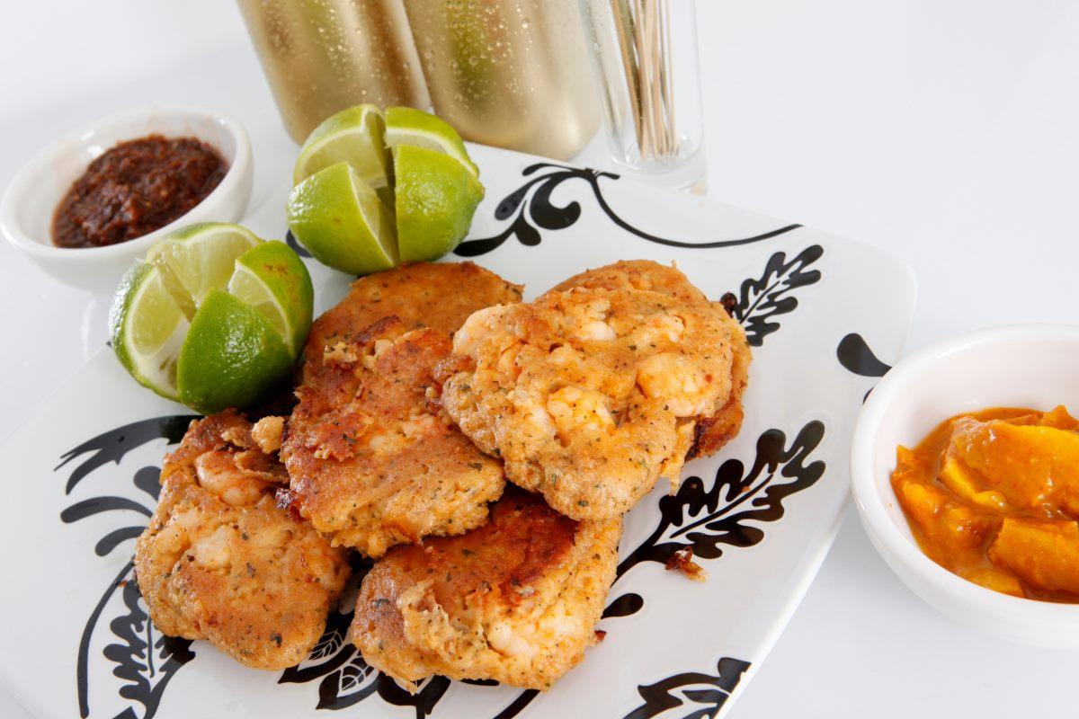Plate of golden-brown seafood fritters served with lime wedges, a spicy dipping sauce, and a tangy chutney on a decorative black-and-white plate with a chilled beverage in the background.