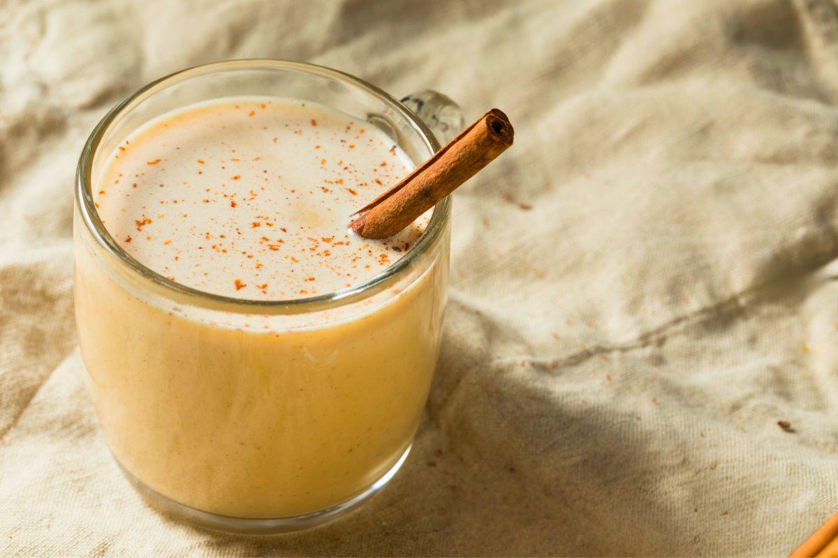 A glass mug filled with creamy eggnog, garnished with a sprinkle of cinnamon or nutmeg and a cinnamon stick, placed on a beige fabric background.