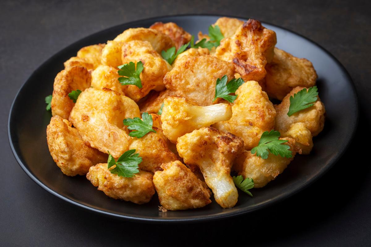 A plate of golden-brown, crispy fried cauliflower florets fried in batter, garnished with fresh parsley leaves, served on a dark plate.