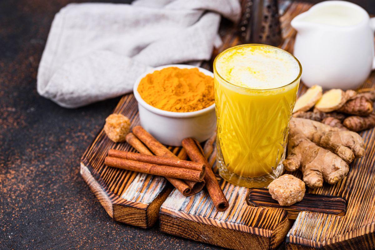 A glass of golden milk surrounded by turmeric powder, ginger root, cinnamon sticks, and a small pitcher of milk on a wooden tray.