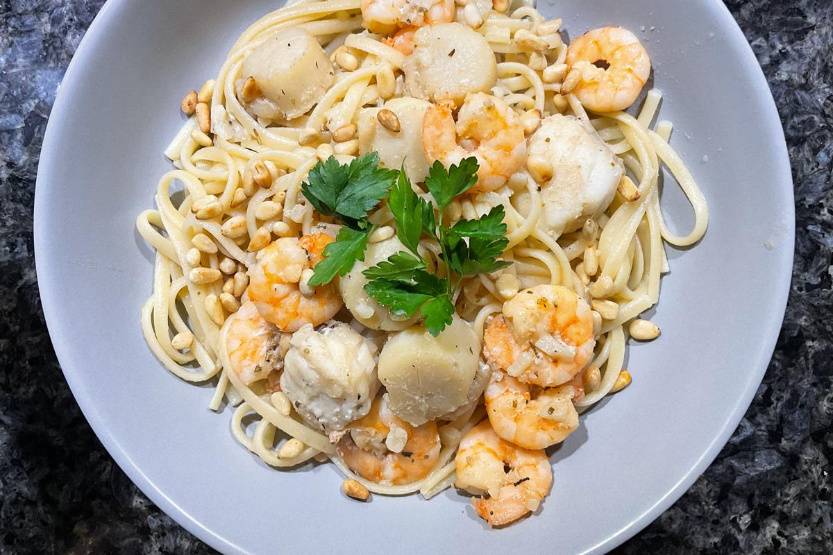 Linguine pasta Freeport, topped with shrimp, scallops, toasted pine nuts, and a sprig of parsley on a gray plate.
