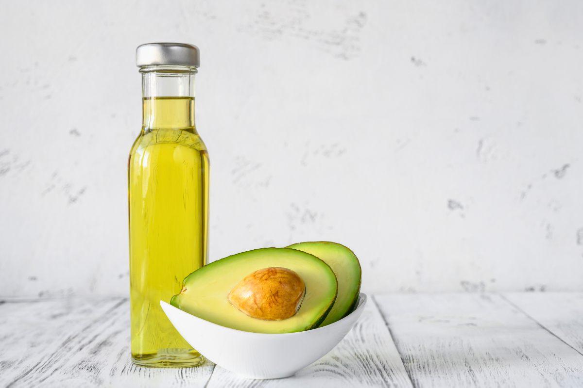 Bottle of avocado oil beside a halved avocado in a white bowl on a white table.