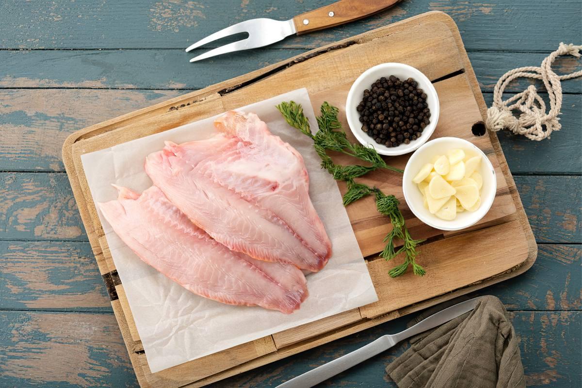 Raw tilapia fillets on wooden board with herbs, peppercorns and sliced garlic by the side.