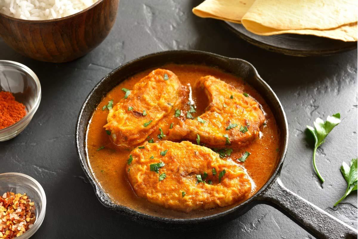 Fish steaks in a rich orange curry sauce in a cast iron skillet, surrounded by bowls of steamed rice, chili powder, chili flakes, and stacked papadums on a dark slate surface.