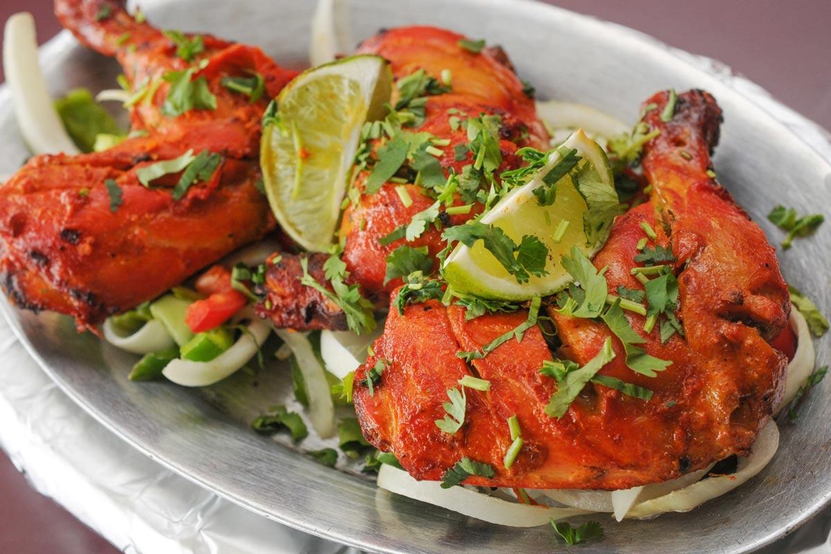 Tandoori chicken pieces with charred red marinade on a foil-lined metal tray, garnished with lime wedges, fresh cilantro, and sliced onions.
