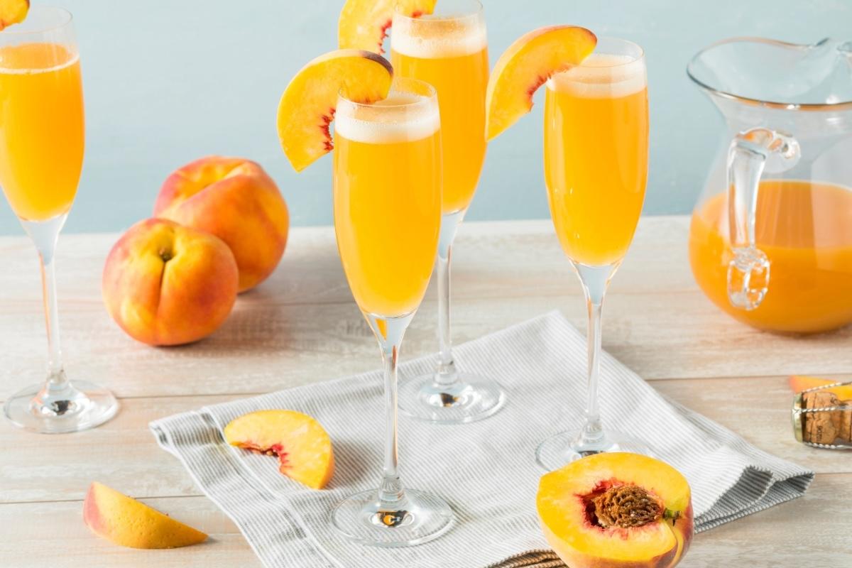 Bellini peach cocktails in champagne flutes with fresh peach slices, surrounded by peaches, a pitcher, and a cork on a light table.