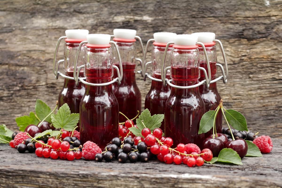 Six small swing-top glass bottles of deep red summer berry cordial on a rustic wooden surface, surrounded by fresh redcurrants, blackcurrants, raspberries, and cherries.