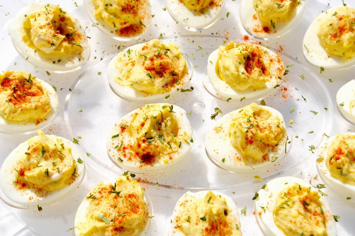 A tray of classic deviled eggs, filled and piped with a creamy yellow yolk mixture, topped with smoked paprika and chopped parsley.