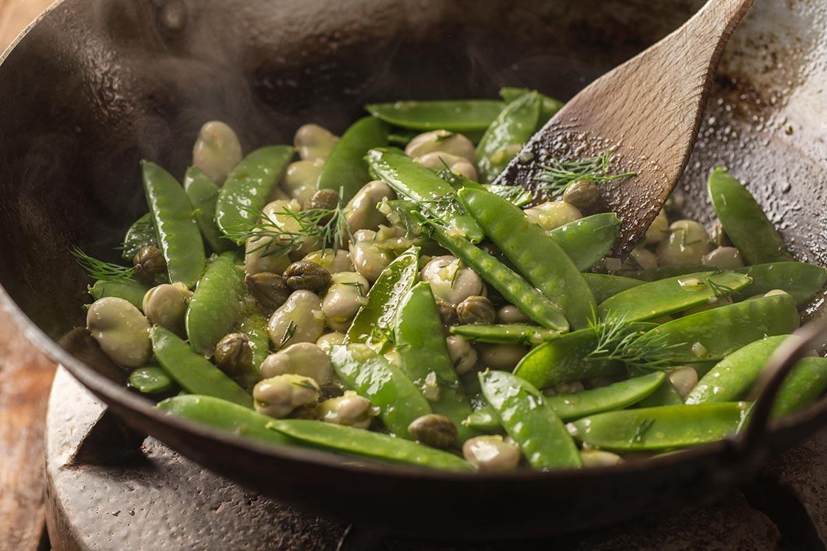 A wok filled with sautéed fava beans, sugar snap peas and capers, garnished with fresh dill, held with a wooden spatula and gently steaming.