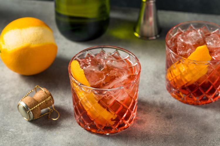 Two glasses of Negroni Sbagliato with orange peels and ice on a grey surface with a peeled orange and bottle stopper nearby.