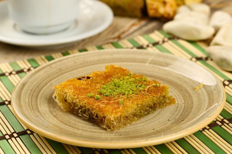 A square piece of kataifi on a beige plate, chopped pistachios. Coffee cup in the background on a bamboo mat.