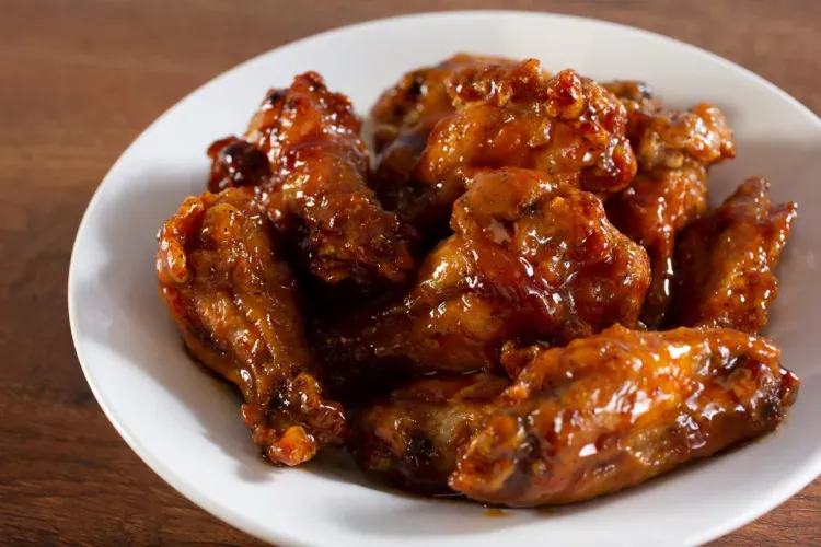 White bowl filled with glazed chicken wings coated in a thick, shiny, reddish-brown sauce, placed on a wooden surface.