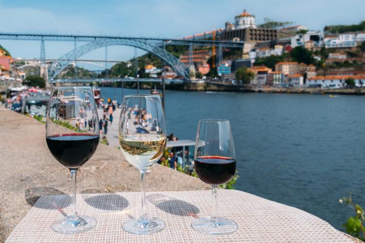 Three glasses of Protuguese wine, red, white, and dark red, on a table overlooking a scenic riverside Proto town with its bridge and colorful buildings on a sunny day.