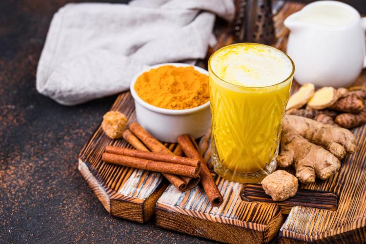 A glass of golden milk surrounded by turmeric powder, ginger root, cinnamon sticks, and a small pitcher of milk on a wooden tray.