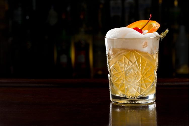 Classic whiskey sour in a patterned glass with a frothy egg white top, garnished with an orange slice and a cherry, set against a dimly lit bar background.
