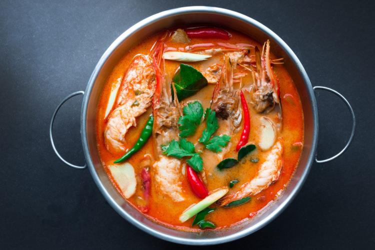 A pot of tom yum goong, Thai spicy and sour shrimp soup with prawns, herbs, and chilies in a bright orange broth.