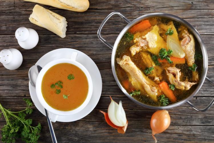 Clear chicken broth in a bowl. A pot with the rest of the broth with the ingredients is on the side.