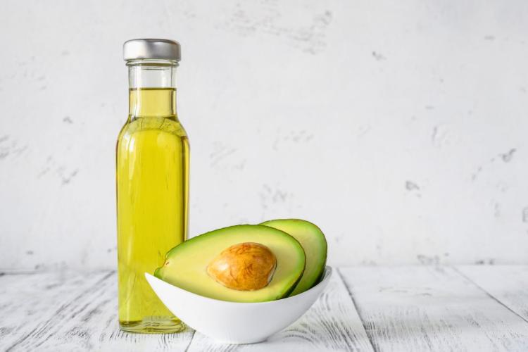 Bottle of avocado oil beside a halved avocado in a white bowl on a white table.