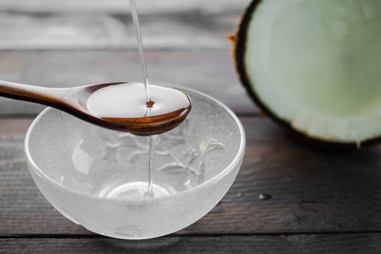 Clear coconut oil pouring onto a wooden spoon over a glass bowl; halved coconut in background.