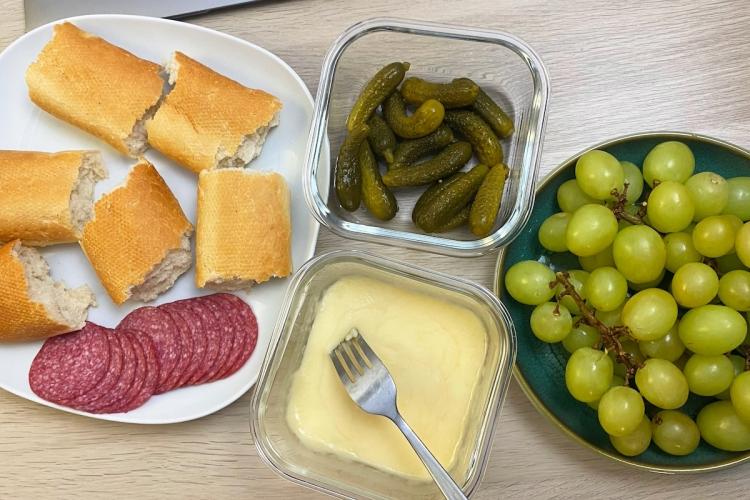 Snack plate with baguette pieces, salami, pickles, melted cheese, and green grapes.