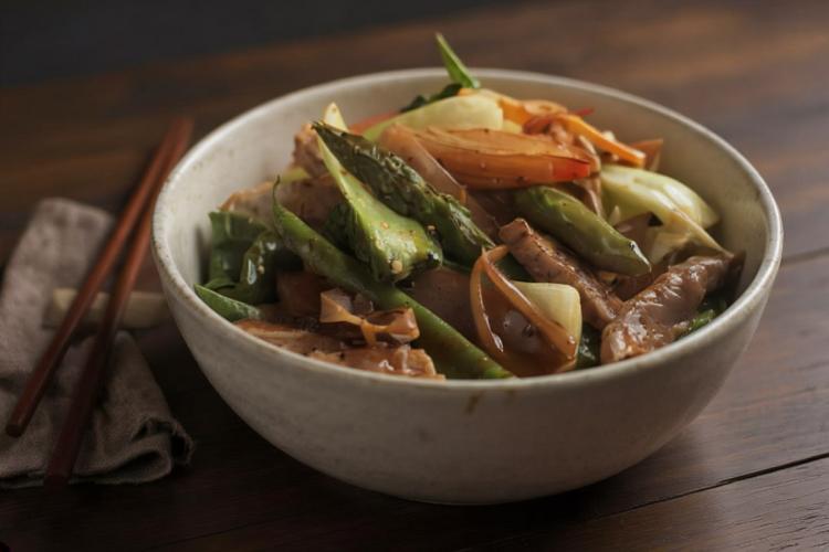 A pale stoneware bowl of pork and vegetable stir-fry — strips of pork with green asparagus, flat green beans, onion wedges, and red bell pepper in a light glossy sauce — on a dark wood table with chopsticks resting on a linen napkin. Synthetic (AI-generated) image.