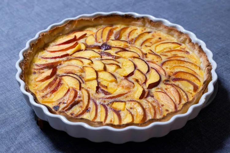 An apple tart in a white fluted ceramic dish, thin apple slices arranged in overlapping rings over a set custard filling, golden and caramelized at the edges, on a blue-gray linen cloth.