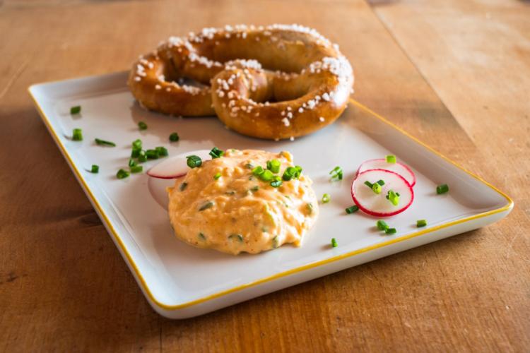 A plate of Obatzda garnished with chopped chives, served alongside a large salted pretzel and thin radish slices.