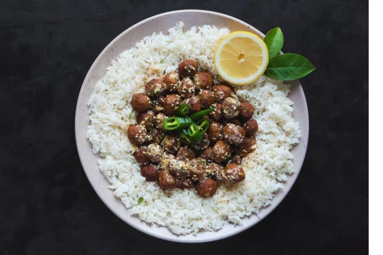 A bowl of Egyptian rice koftas (koftet rozz) — small fried beef-and-rice meatballs sprinkled with sesame seeds — arranged in a ring of white rice and topped with sliced green chile and half a lemon, on a dark background.