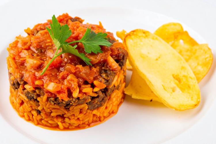 A molded round of spiced tomato-red rice topped with a savory sauce and a flat-leaf parsley sprig, with two golden fried potato slices alongside, on a white plate.