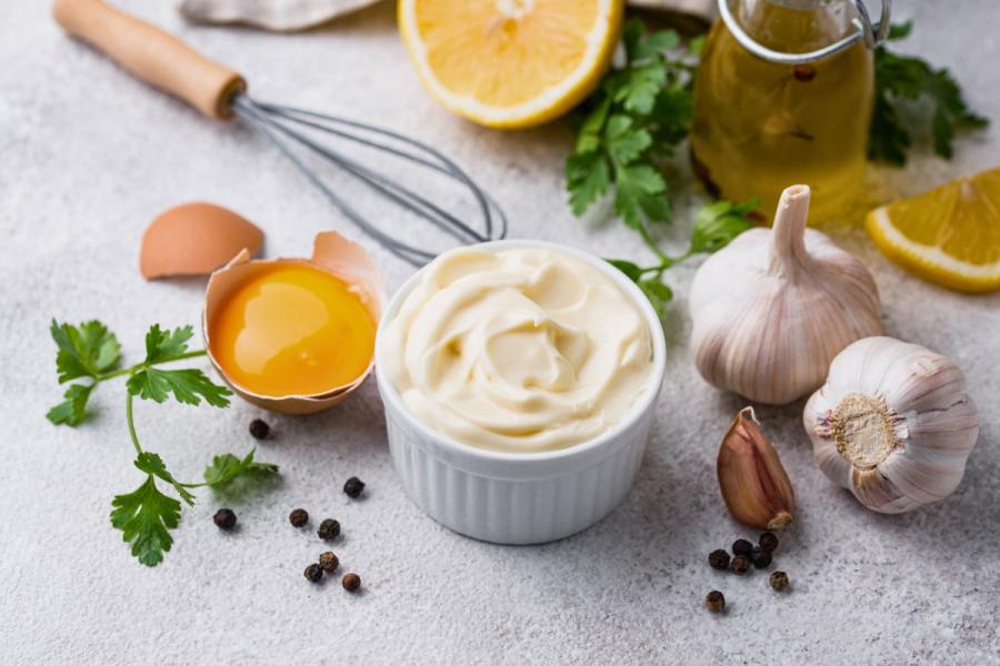 Ramekin of creamy mayonnaise surrounded by a cracked egg, garlic, parsley, black peppercorns, lemon slices, and a whisk.