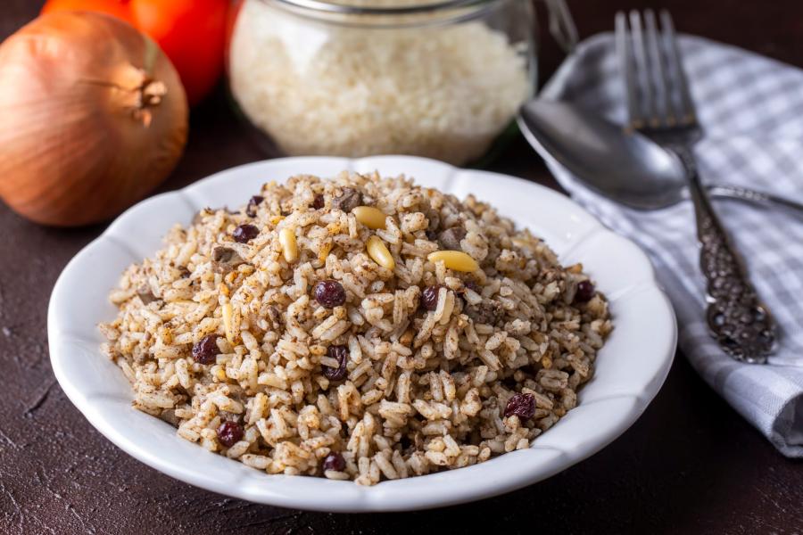 A bowl of pilaf rice topped with pine nuts and currants, served in a white bowl. In the background, there are an onion, a jar of rice, and tomatoes, with a fork and spoon on a gray checkered napkin.