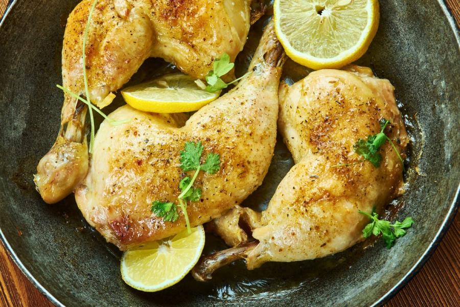 Close-up of four sumac-seasoned roasted chicken legs in a pan, garnished with fresh herbs and lemon wedges.
