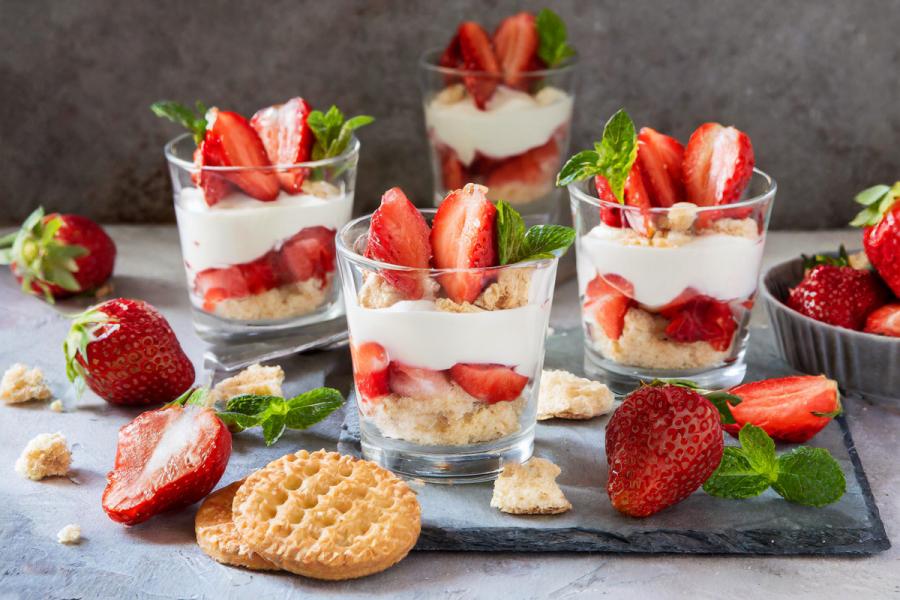 Strawberry cheesecake parfait cups with layers of crumbled cookies, fresh strawberries, and creamy yogurt-cheese mix, garnished with mint leaves, surrounded by freshl strawberries and cookie pieces.
