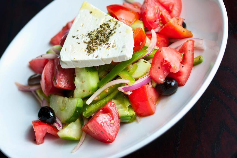 A fresh Greek salad with tomatoes, cucumbers, green bell peppers, red onions, black olives, and a slice of feta cheese sprinkled with oregano on a white plate.