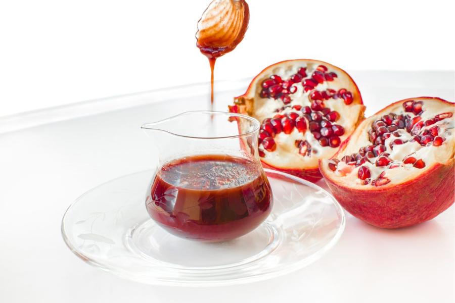 Glass jug filled with deep red pomegranate sauce on a decorative glass plate, accompanied by fresh pomegranate halves and a honey dipper dripping the sauce.