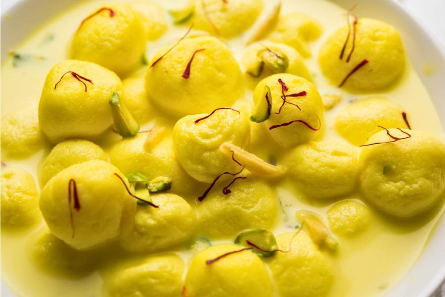 Close-up of Rasmalai, featuring yellow paneer balls soaked in saffron-infused milk, garnished with slivers of pistachios, almonds, and saffron strands.