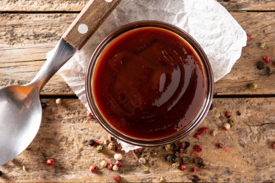 Close-up of a glass container filled with glossy barbecue sauce on a wooden surface, surrounded by scattered peppercorns and a spoon with a wooden handle.