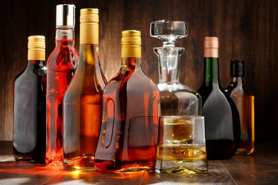 An assortment of spirit bottles in various shapes and colors on a wooden surface, with a glass of whiskey and a decanter.