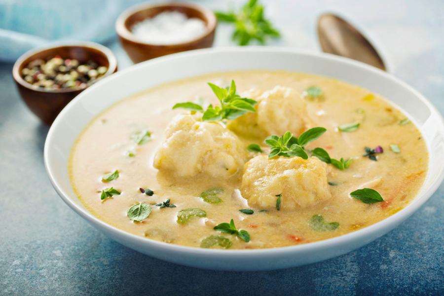 A bowl of creamy chicken soup with fluffy dumplings, garnished with fresh herbs, served on a blue surface with seasoning bowls and a wooden spoon in the background.
