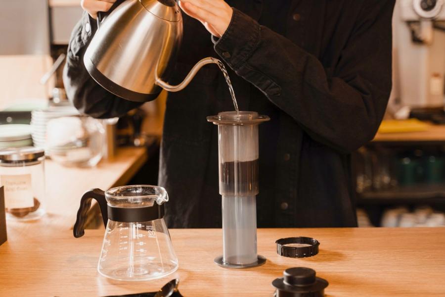 Person pouring hot water from a stainless steel gooseneck kettle into an AeroPress coffee maker on a wooden countertop, with a clear glass coffee server and AeroPress accessories nearby.