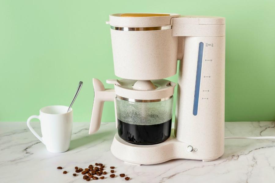 Light beige automatic drip coffee maker with glass carafe filled with black coffee on marble countertop, white ceramic mug with spoon beside it, and scattered coffee beans, set against a green background.