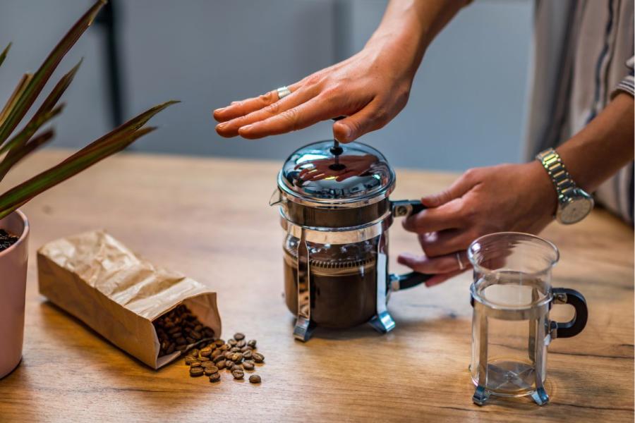 Person pressing plunger on French press coffee maker on wooden table, with empty glass beaker, spilled coffee beans from paper bag, and potted plant nearby.
