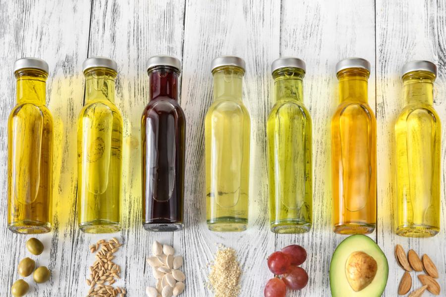 A row of different cooking oils in glass bottles with their source ingredients like olives, seeds, and avocado on a white wood background.