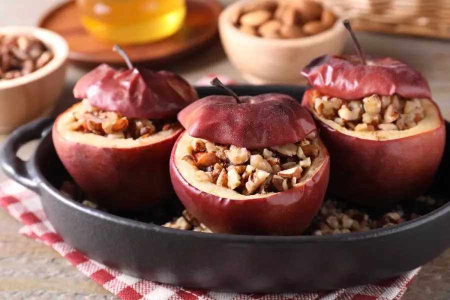 Three red baked apples stuffed with chopped nuts in a black baking dish, sitting on a red-and-white checkered cloth with bowls of nuts and honey in the background.